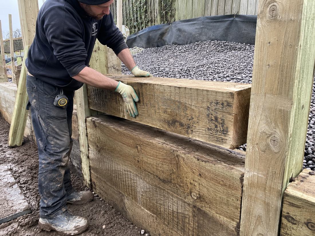 Builder placing a treated timber sleeper into a retaining wall with drainage metal and geotextile visible behind